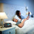 A man wearing a sleep mask lies in bed using a sleep apnea device on a bedside table, while a woman sits up beside him reading a book in a softly lit bedroom.